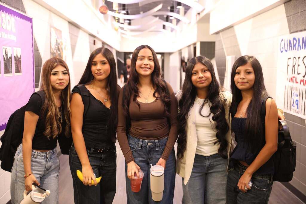 Five high school girls standing together in a school hallway, smiling as they pose for a group photo. They are casually dressed in jeans and neutral-colored tops, carrying backpacks, water tumblers, and snacks.