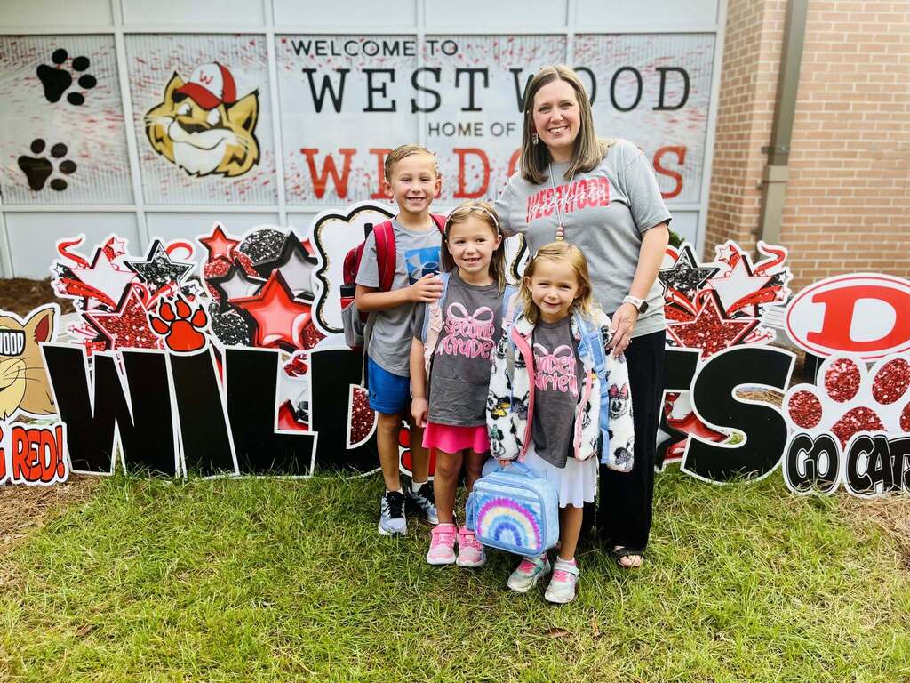 A smiling woman stands outside with three young children in front of a decorative school photo backdrop that reads "WELCOME TO WESTWOOD HOME OF WILDCATS." The young boy and two young girls wear school shirts and backpacks, and the youngest holds a colorful tie-dye lunchbox.