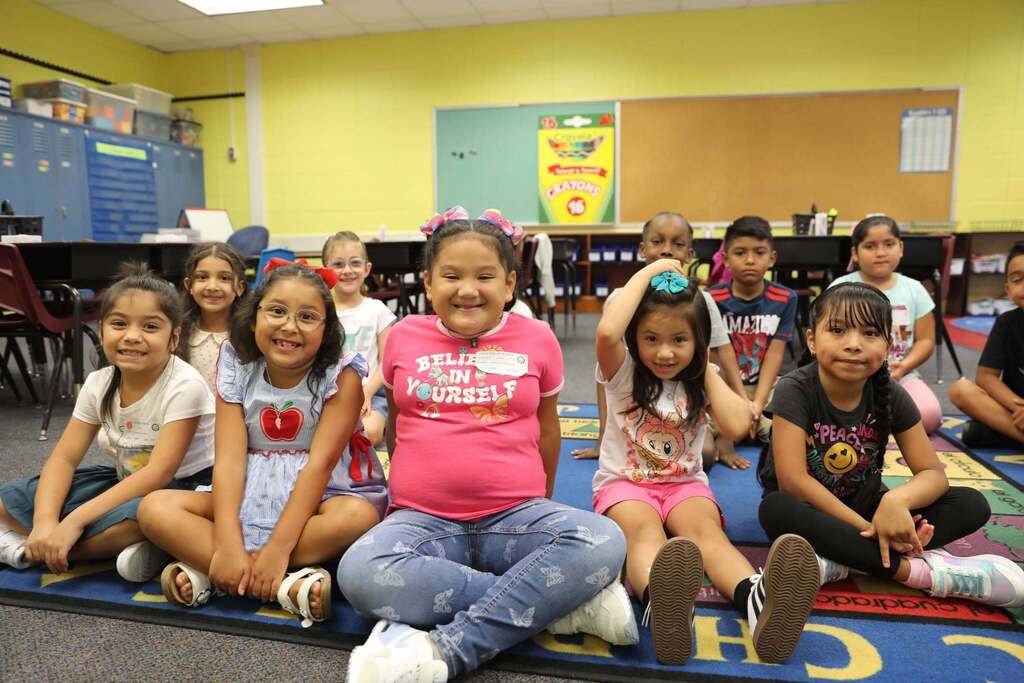 A group of elementary school children sitting cross-legged on a colorful alphabet rug in a bright classroom, smiling happily at the camera on their first day of school.