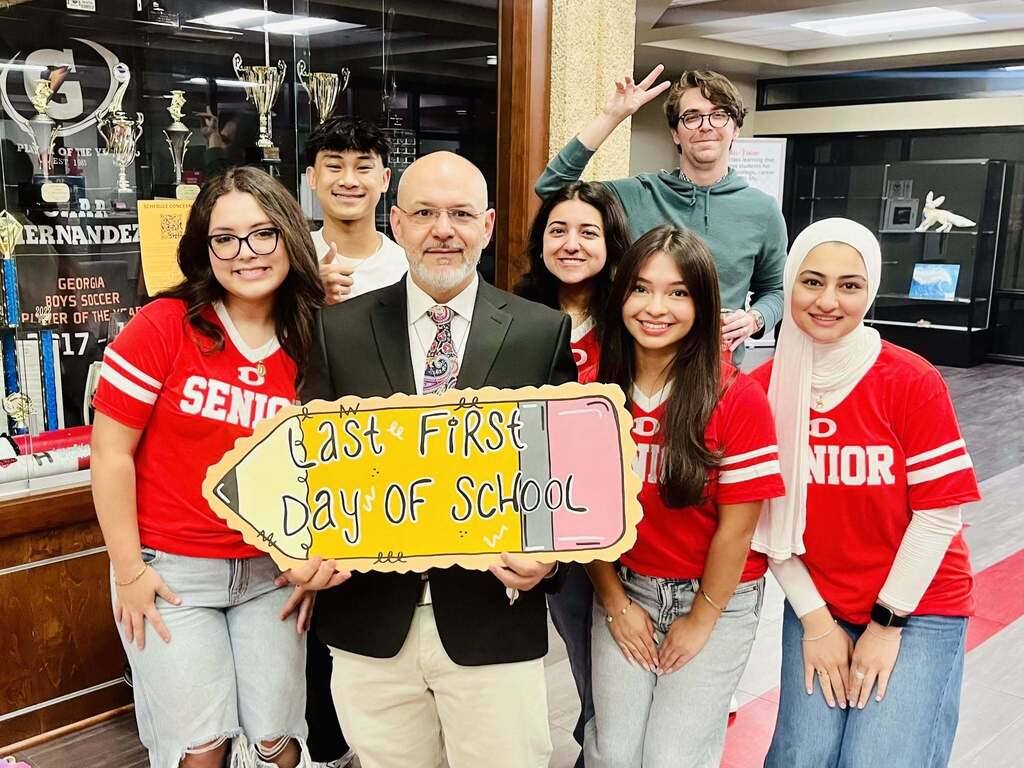 A male school administrator in a suit and tie standing with a group of high school seniors wearing red "SENIOR" t-shirts. Together, they hold a large pencil-shaped sign that reads "Last First Day of School" in front of a trophy display case.