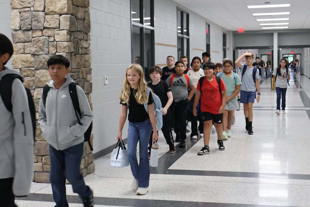 A line of middle school students carrying backpacks, lunch bags, and water bottles walking down a bright, modern school hallway with stone accents and tile floors on their way to class.