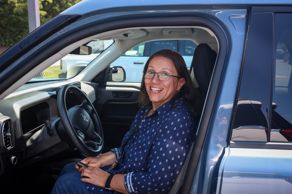 A smiling woman with dark hair and glasses sits in the driver's seat of a light blue Ford vehicle with the driver-side door open. She wears a blue pattern button-down shirt and holds a key fob in her hands while looking back toward the camera.