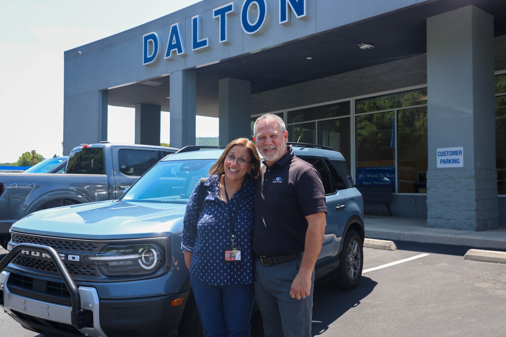 A smiling woman with glasses in a blue patterned button-down shirt stands beside a male dealership representative in a black polo shirt. They are posing in front of a light blue Ford Bronco Sport parked in the lot directly outside a dealership building with large blue "DALTON" signage overhead.