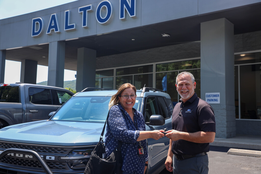 A male dealership representative in a black polo shirt hands a key fob to a smiling woman in a blue patterned shirt. They stand together in front of a light blue Ford Bronco Sport in the parking lot outside a dealership building with large blue "DALTON" lettering overhead.
