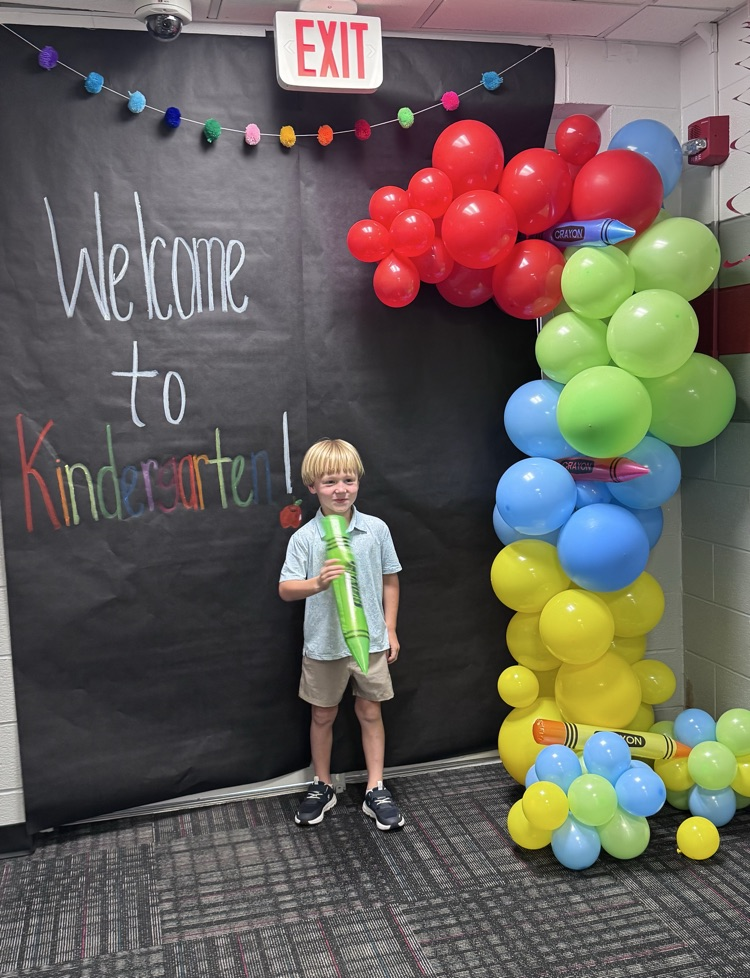Student holding a big inflatable crayon standing in front of a welcome to kindergarten sign during the meet the teacher event 