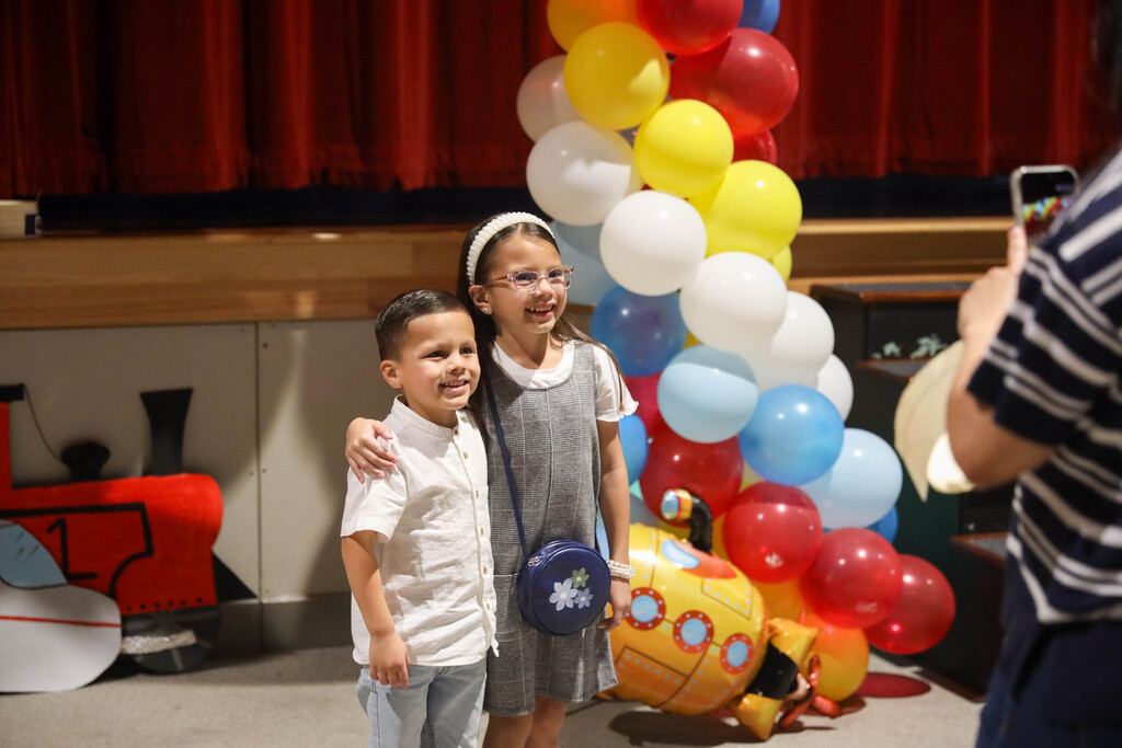 A young boy in a white short-sleeved button-up shirt and light jeans stands next to a young girl wearing a grey plaid jumper, headband, glasses, and a blue crossbody purse. They smile together in front of a colorful balloon arch and red stage backdrop while a person standing on the right holds up a smartphone to photograph them.