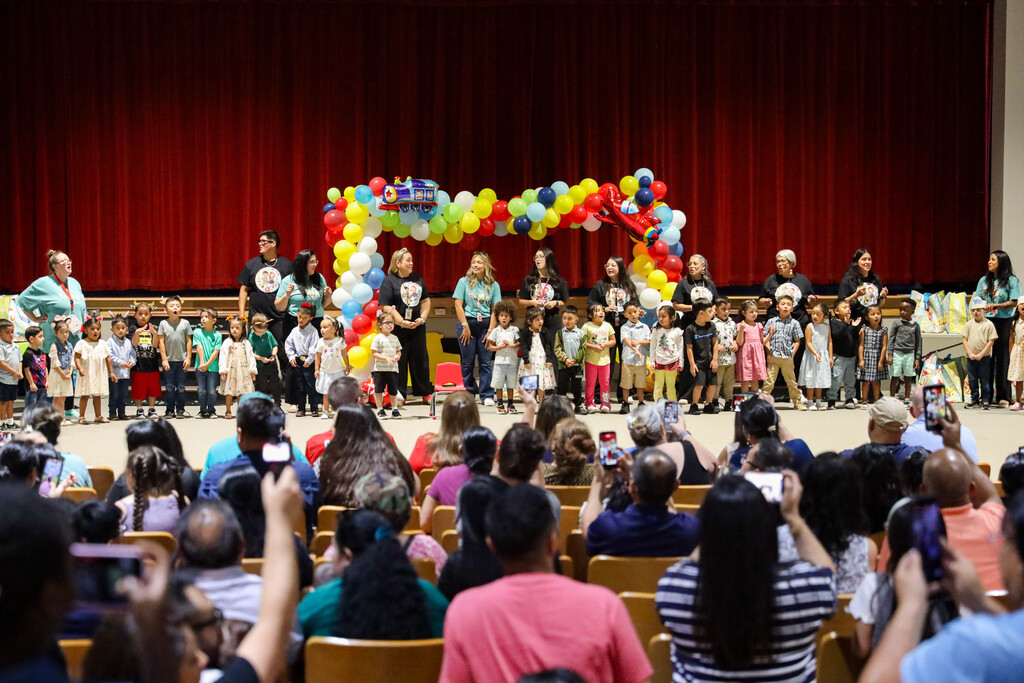 An auditorium audience viewed from behind holds up smartphones to take photos of a large group of young children and staff lined up on stage. The stage features a colorful balloon arch in the background against dark red curtains.