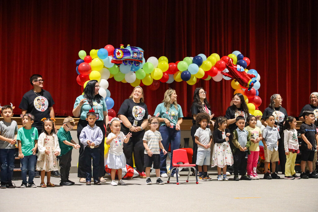 A line of young children wearing paper headbands stands at the front of a stage during an event. Behind them, teachers and staff stand smiling under a large arch made of red, yellow, green, and blue balloons with vehicle-themed decorations, set against a red stage curtain.
