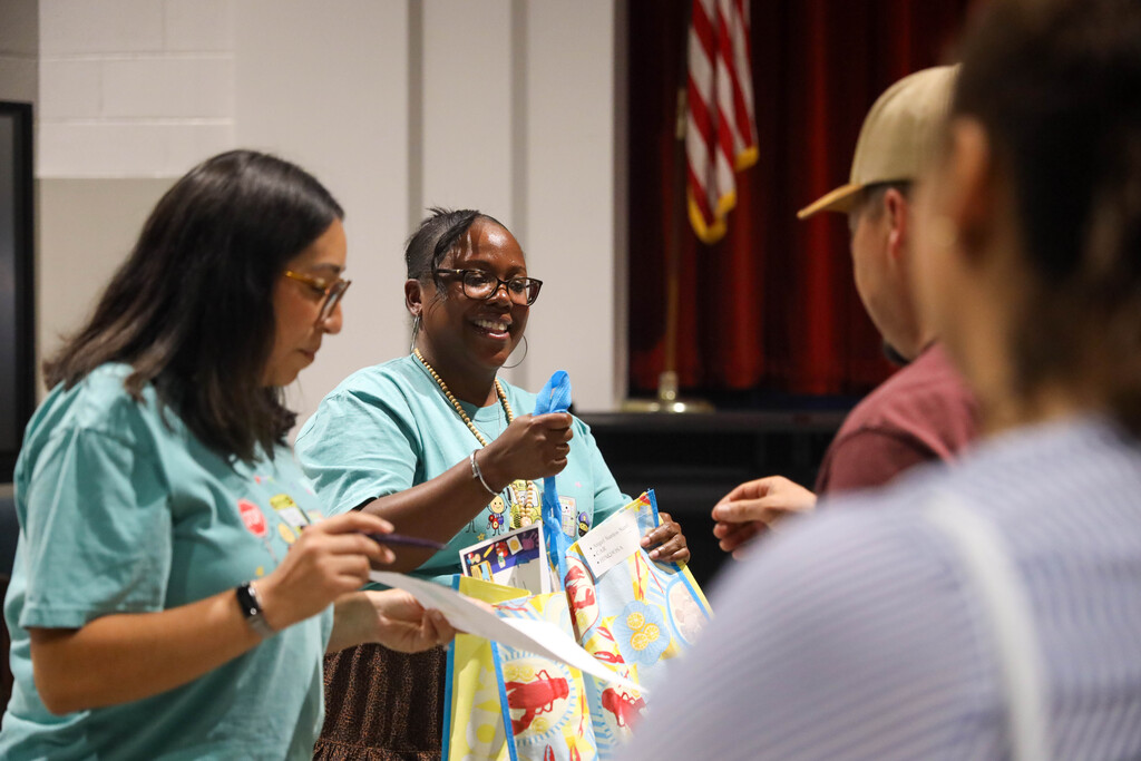 Two female staff members wearing teal t-shirts interact with parents in an auditorium setting. The woman in glasses on the left reviews paperwork, while the smiling woman in the middle hands a blue lanyard and a colorful gift bag containing learning materials to a parent standing in the foreground.
