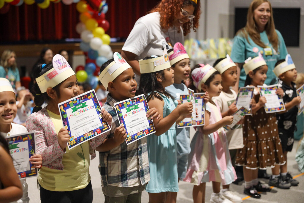 A line of smiling young students wearing paper crowns stand side-by-side on stage, holding up their Pre-Kindergarten completion certificates facing forward. Teachers stand behind them encouraging the children as parents watch from the side.