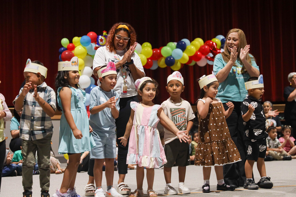 A row of young children wearing hand-decorated paper crowns stand on stage making hand gestures during a performance. Behind them, two female teachers cheer and clap along in front of a colorful balloon display and red stage curtain.