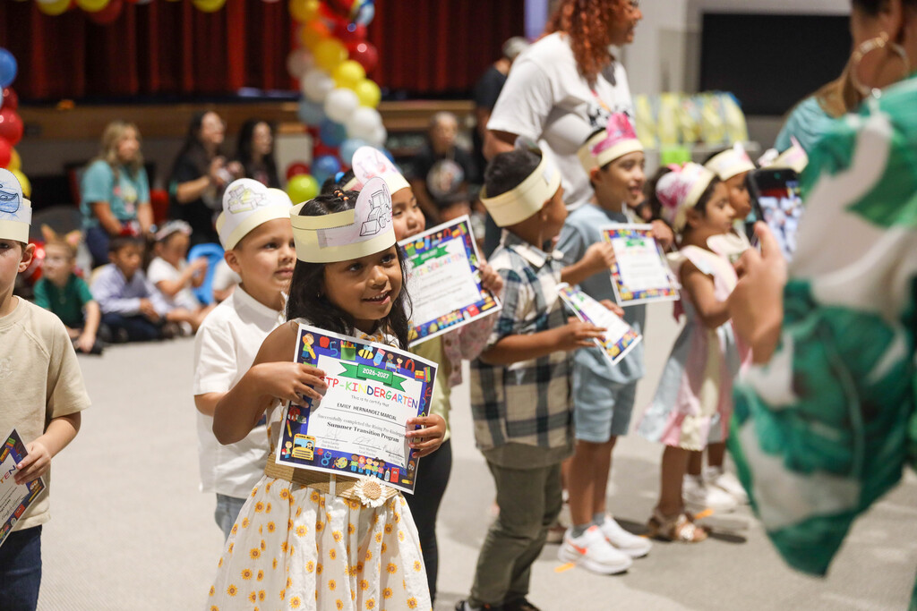 A line of young students wearing decorated paper crowns walk in sequence across an auditorium stage. Each child proudly holds a colorful certificate for completing the Pre-Kindergarten Summer Transition Program while teachers guide them from behind.