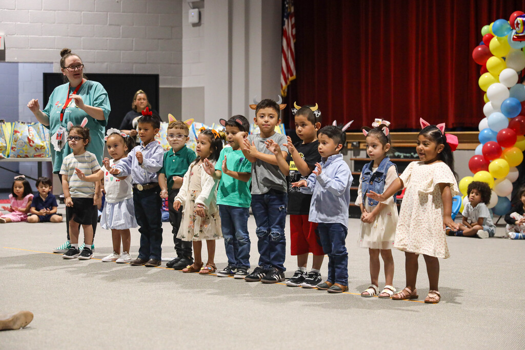 A row of young students wearing headband-mounted paper animal ears stand in a line on stage, raising their hands to follow motions. On the far left, a female teacher in a teal shirt and lanyard demonstrates the movements for the children to mirror.