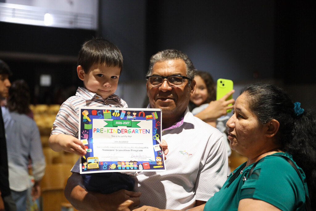 A young boy held by an older man holds up a colorful Pre-Kindergarten Summer Transition Program certificate reading "2026-2027 Pre-Kindergarten". A smiling woman in a green top stands beside them, while another person in the background captures the moment on a bright yellow smartphone.