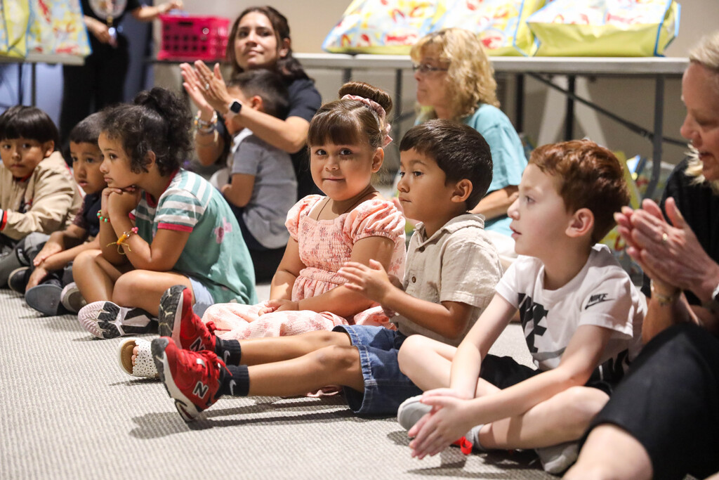 A group of young children sit cross-legged in a row on a carpeted floor during an indoor event. A young girl in a pink dress in the center smiles toward the camera while surrounding children look toward the front and adults nearby clap.
