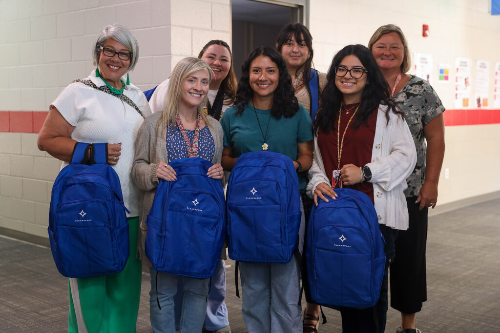 Six female staff members stand close together in a cinderblock school hallway, posing with royal blue CleanSpark-branded backpacks. Four of the women in the front hold the backpacks forward, while the others smile from behind in casual professional attire.