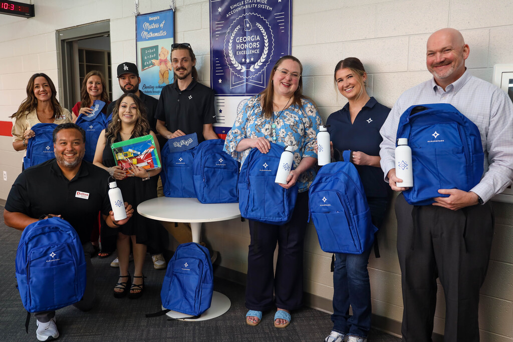 Eight staff members pose for a group photo in a school corridor beside a white round table loaded with blue backpacks. The individuals hold up blue CleanSpark backpacks, white insulated water bottles, and clear pencil pouches filled with school supplies while smiling at the camera.