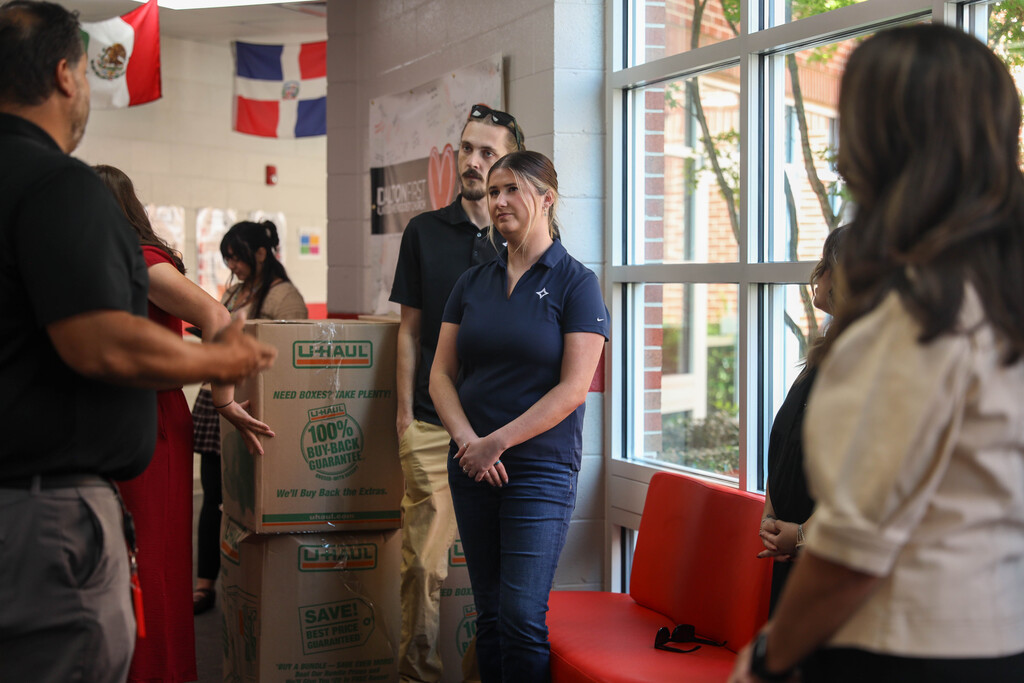 A woman in a navy CleanSpark polo shirt and jeans stands with hands clasped in a school hallway, listening to a colleague off-camera on the left. Behind her, stacked U-Haul boxes rest against the wall under hanging international flags.