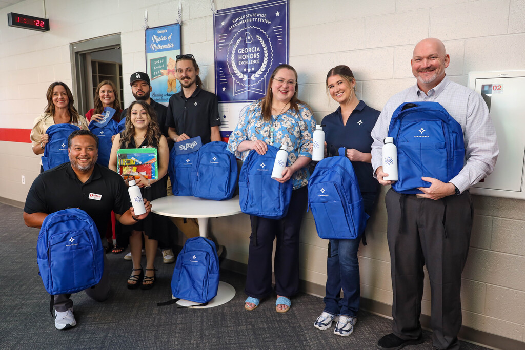 Eight adults pose together around a white pedestal table in a school hallway beneath educational posters. One man kneels in front holding a blue backpack and a white CleanSpark water bottle, while others stand holding matching backpacks, water bottles, and school supply pouches.