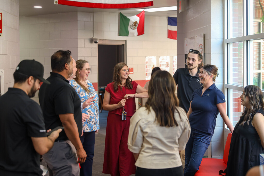 A group of eight adults stand in a circle near a large windowed exterior wall in a school hallway, engaged in conversation. Mexican and Dominican flags hang suspended from the ceiling in the background, along with stacked U-Haul cardboard boxes along the wall.