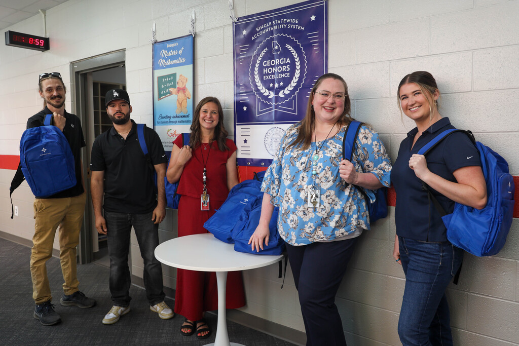 Five school staff members pose along a hallway wall under a blue "Georgia Honors Excellence" banner. Three women and two men hold or wear blue CleanSpark backpacks while smiling for the photo around a small white table.
