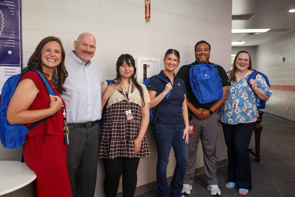 Six individuals stand in a row along a white cinderblock hallway wall inside a school building. A woman in red on the far left wears a blue CleanSpark backpack, a man in the center holds one up to his chest, and others wear or hold matching backpacks while smiling toward the camera.