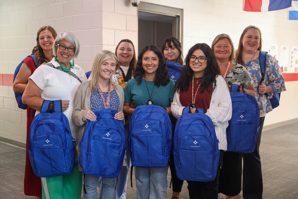 Eight female school staff members pose together in a brightly lit hallway. Five women in the front row hold royal blue CleanSpark backpacks, while three colleagues stand behind them smiling. Flags from different countries hang along the ceiling beam above.