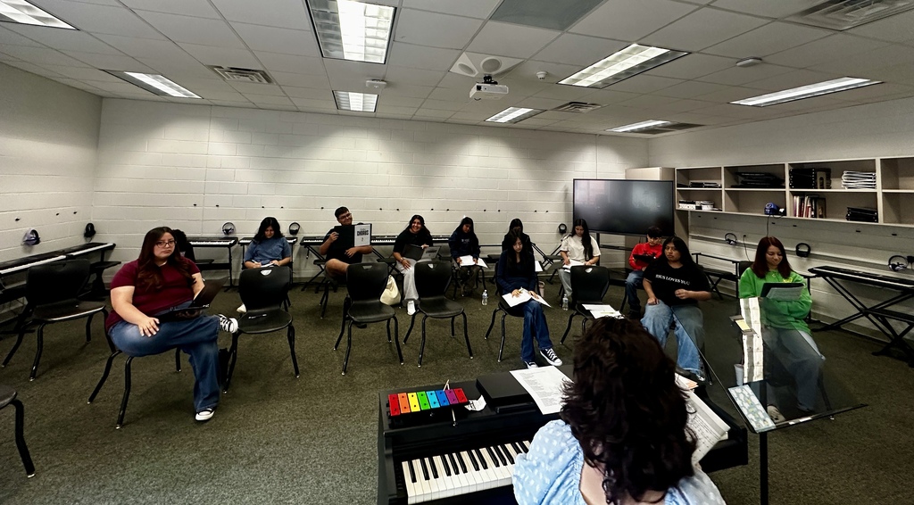 View from behind an instructor seated at an electric keyboard as high school chorus students sit in a wide semi-circle around the room holding sheet music folders during a music lesson.