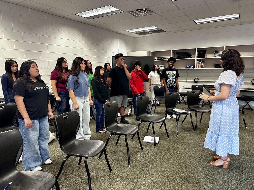 A group of high school students stands in a semicircle inside a music classroom, rehearsing under the direction of new chorus teacher Bryanna Cali, who wears a blue polka-dot dress and gestures as she leads the class behind rows of black chairs.