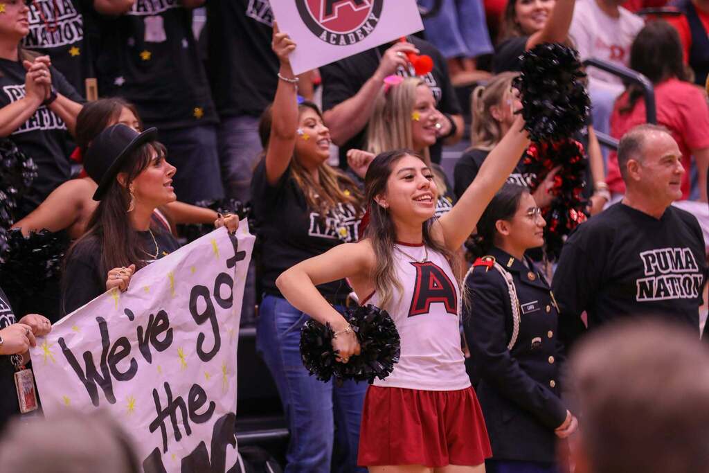 A cheerleader in a white tank top with a red "A" logo and red shorts smiles while raising a black pom-pom in a crowded gymnasium bleacher section. Beside her, a woman wearing a black fedora holds a handmade banner reading "We've got the magic", while other fans in black "PUMA NATION" shirts cheer, wave signs, and raise pom-poms in the background.