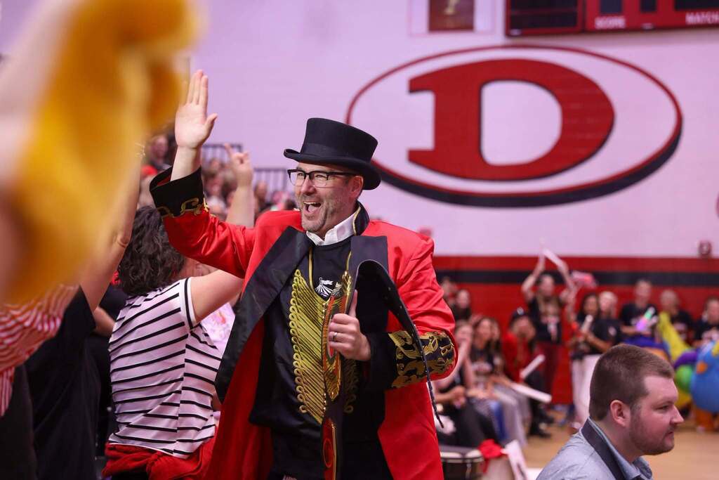 Principal Brandon Headrick dressed as a ringmaster in a black top hat, glasses, and an ornate red coat with gold embroidery high-fives Park Creek staff  on a gymnasium floor. He smiles broadly while carrying a large championship belt under his arm, with a large red "D" logo visible on the gym wall behind him.
