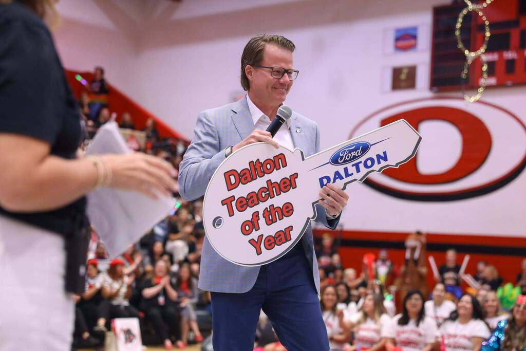 Jason Denson, the owner of Ford of Dalton, in a blue plaid suit coat, glasses, and blue trousers smiles as he speaks into a microphone while holding a large novelty key in a crowded gymnasium. The key features the Ford logo and reads "Dalton Teacher of the Year" in red outline lettering. In the background, audience members cheer from the bleachers beneath a large red "D" logo on the wall.