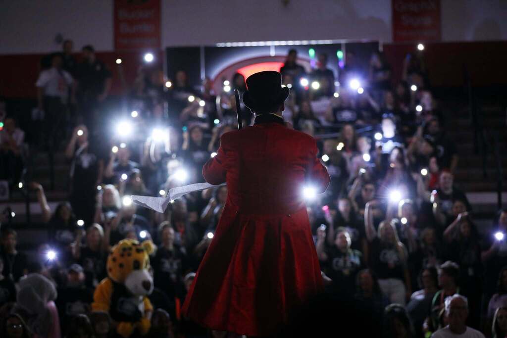 The Park Creek Elementary School principal Brandon Headrick seen from behind in a red tailcoat stands on stage before a large crowd in a dark arena, with audience members holding up lit phone flashlights and the park creek mascot  in the background.