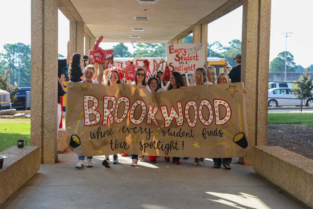 A enthusiastic group of teachers and staff walk down an outdoor covered walkway holding school spirit signs. In front, several women hold a large brown paper banner that reads "BROOKWOOD where every student finds their spotlight!" in red marque-style lettering and black script. Behind them, staff members wave red foam fingers, pom-poms, and hold signs including one that reads "Every Student HAS A Spotlight!".