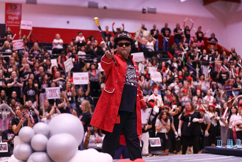 Deputy Superintendent  Nick Sun in a bright red, gold-trimmed coat, top hat, and sunglasses points a cane directly toward the camera inside a crowded gymnasium. He wears a black shirt that reads "THE GREATEST SHOW IN EDUCATION" and carries a clipboard. Behind him, packed bleachers full of cheering teachers and staff hold signs, glow sticks, and pom-poms under red arena lighting.