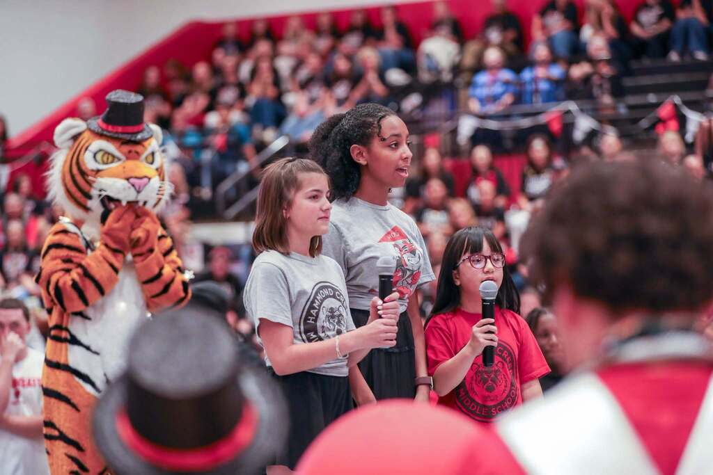 Three young students stand on a gym floor singing into microphones during an assembly. Two are wearing grey Hammond Creek t-shirts, and one in red glasses wears a red Hammond Creek Middle School shirt. To the left, the Hammond Creek  tiger mascot wearing a small black top hat clasps its paws together with educator-packed bleachers visible in the background.