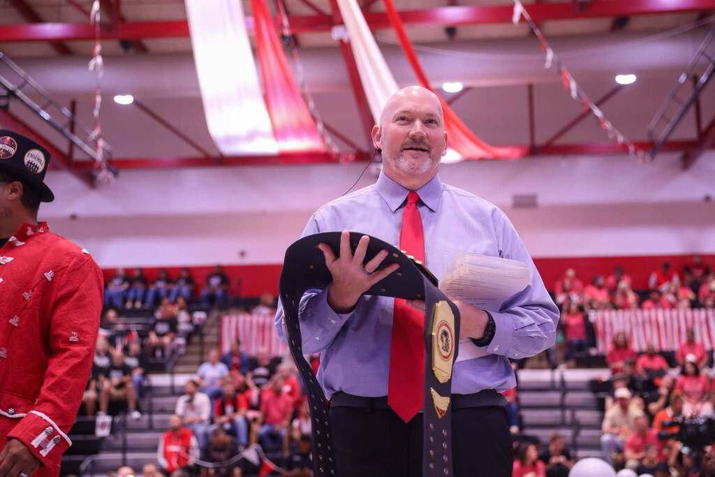 Superindendent Dr. Steven Craft in a light blue dress shirt, bright red tie, and headset microphone presents a large championship belt on a gymnasium floor. He holds papers in one hand, while red and white streamers hang from the ceiling above and audience members fill the bleachers in the background.