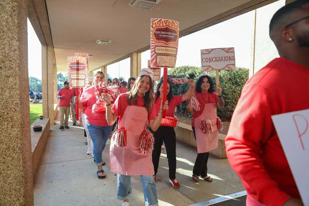 A group of smiling educators dressed as carnival concessions workers walk in a line down an outdoor covered walkway. They wear red shirts, red-and-white striped aprons, paper diner hats, and carry pom-poms along with signs reading "CONCESSIONS" and "THE GREATEST SHOW IN EDUCATION."