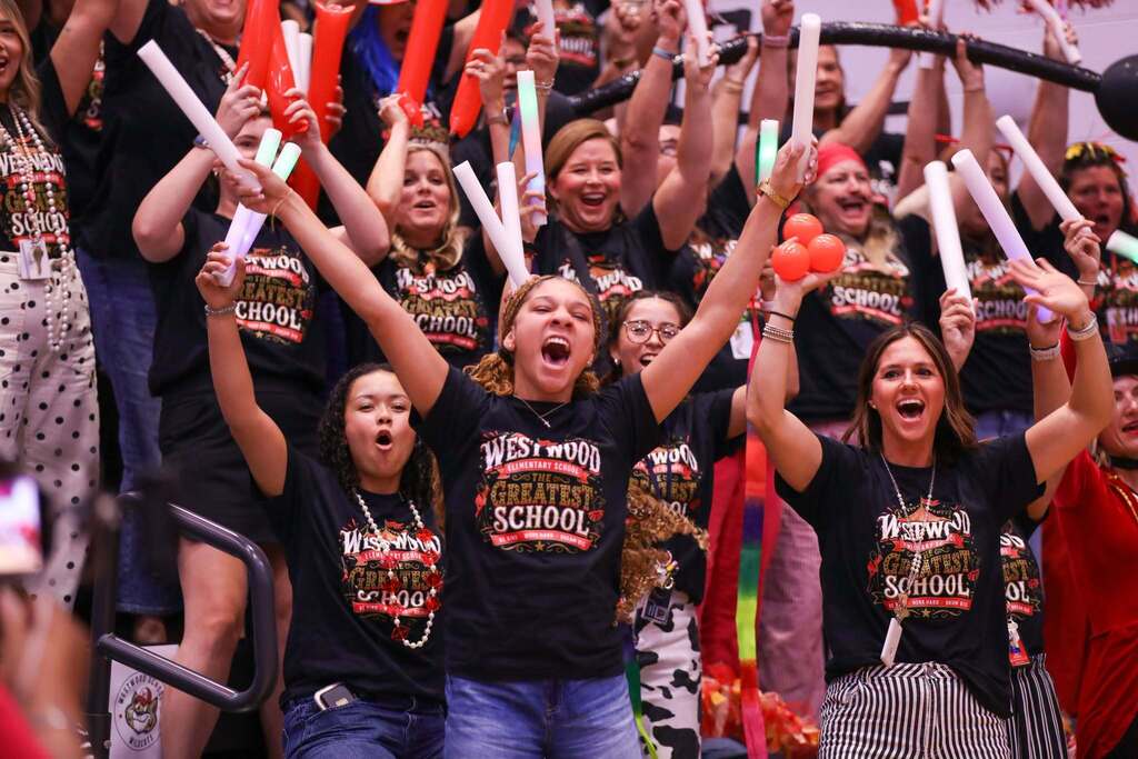A high-energy crowd wearing matching black t-shirts that read "WESTWOOD ELEMENTARY SCHOOL THE GREATEST SCHOOL" cheers enthusiastically in bleachers. In the foreground, three women raise their arms with wide smiles and excited expressions while holding white foam light sticks and orange props. Surrounding fans cheer along, waving red inflatable thundersticks and light-up batons.