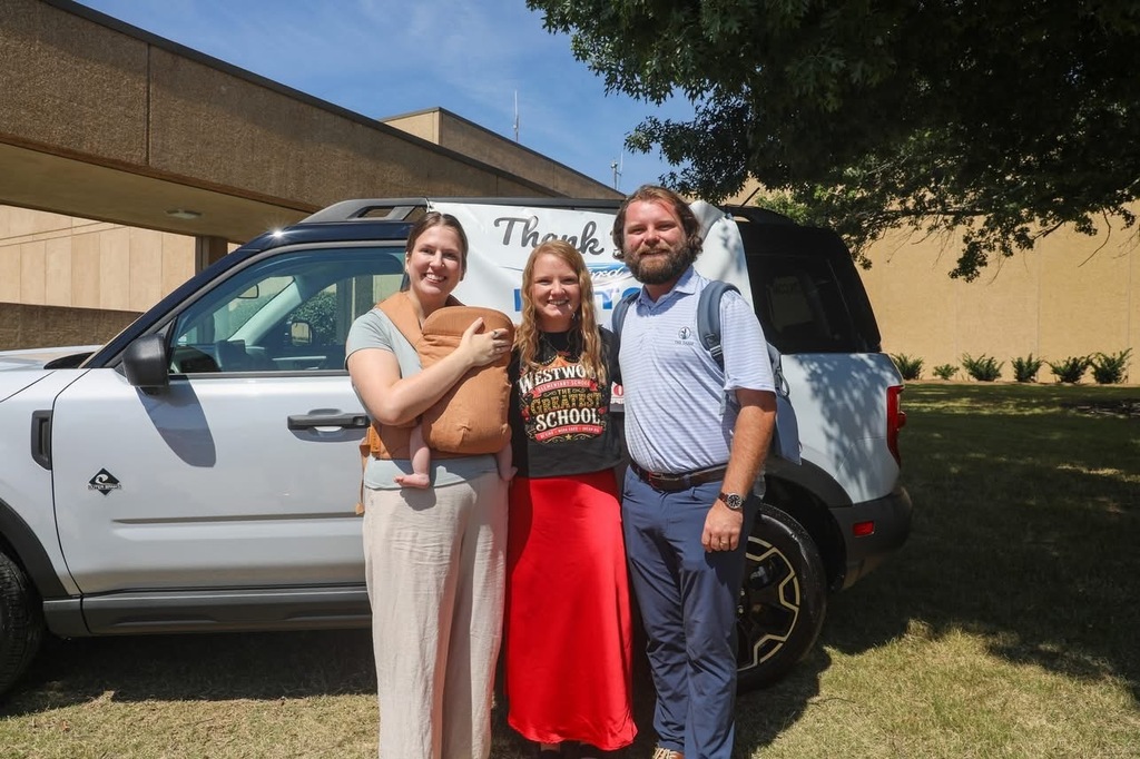 Three adults stand together outdoors smiling in front of a white Ford SUV. The woman in the middle wears a black "WESTWOOD ELEMENTARY SCHOOL THE GREATEST SCHOOL" shirt and a long red skirt. On her left, a woman holds a baby in a beige front carrier, and on her right, a bearded man in a light blue striped polo and navy trousers stands with them on a grassy lawn.