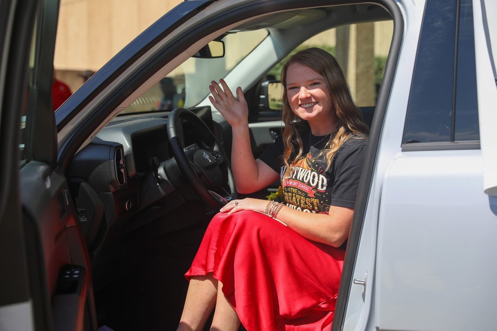 A smiling woman wearing a red skirt and a black "WESTWOOD ELEMENTARY SCHOOL" t-shirt sits in the driver's seat of a white vehicle with the door open, waving at the camera.