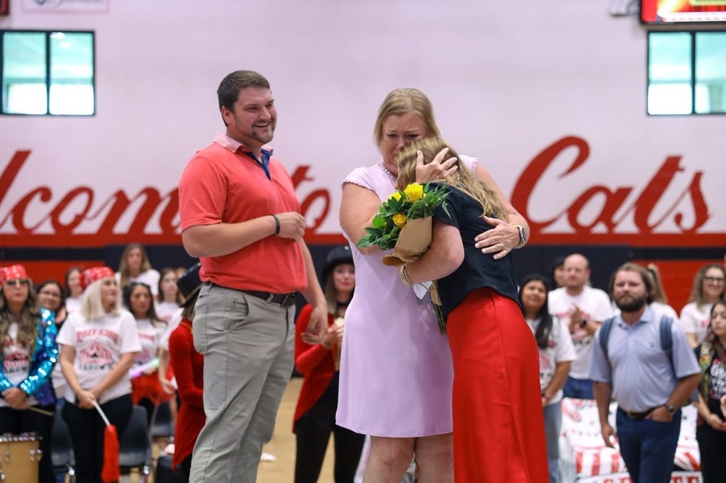 A woman in a light pink dress emotionally embraces a teacher in a red skirt who is holding a bouquet of yellow roses on a gymnasium floor. Beside them, a smiling man in a red polo shirt watches the embrace, while other attendees look on in the background against a wall displaying the words "Welcome Cats".