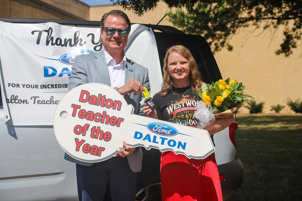 A man in sunglasses and a blue plaid suit coat hands a car key fob to a smiling female teacher standing outdoors in front of a white Ford SUV. Together, they hold a large novelty key reading "Dalton Teacher of the Year" and "Ford DALTON." The teacher wears a red skirt and black Westwood Elementary shirt while holding a bouquet of yellow roses in her other arm.