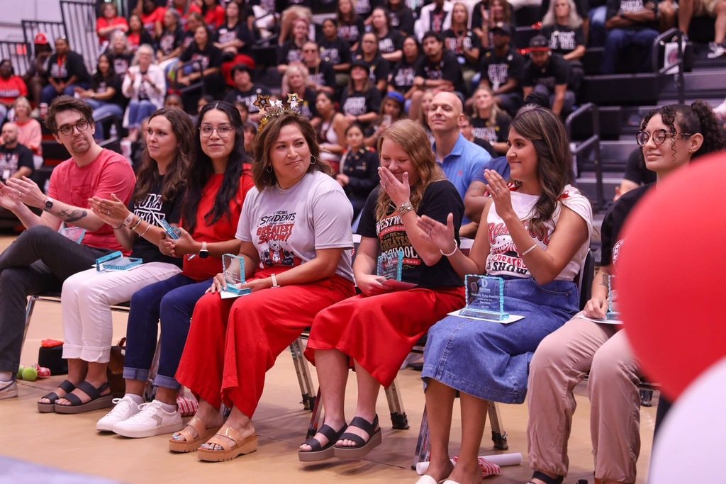 A row of teachers seated on chairs on a gymnasium floor applaud during an awards ceremony. They hold acrylic award plaques in their laps. Near the middle, a female teacher wearing a star headband and red pants smiles towards her colleague in a black Westwood Elementary shirt and red skirt, who covers her mouth in surprise and emotion. In the background, audience members watch from packed bleachers.