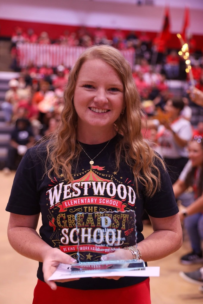 A smiling blonde female teacher stands in a gymnasium holding an engraved glass award plaque. She wears a black t-shirt that reads "WESTWOOD ELEMENTARY SCHOOL THE GREATEST SCHOOL" and a red skirt, with spectators in red and black seated in the blurred bleachers behind her.