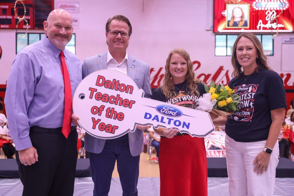 Four smiling people pose together on a gymnasium stage for an award presentation. Two men in suits and a woman on the right present a large novelty car key reading "Dalton Teacher of the Year" and "Ford DALTON" to a female teacher in a red skirt who holds a bouquet of yellow roses.
