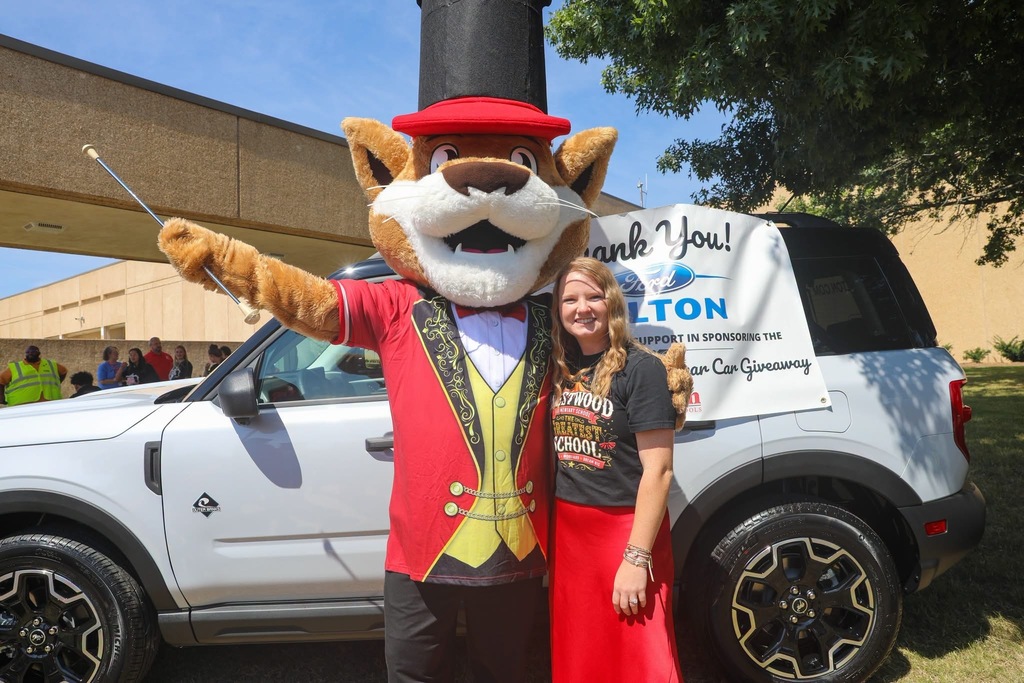 A smiling woman in a red skirt and black Westwood Elementary School shirt poses next to a feline mascot dressed as a ringmaster in a red jacket, yellow vest, and black top hat. The mascot holds out a baton in front of a white Ford SUV displaying a banner reading "Thank You! Ford DALTON... Sponsoring the Teacher of the Year Car Giveaway."