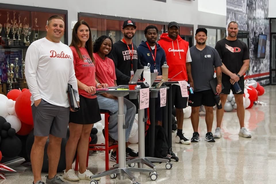 A group of seven smiling adults standing side-by-side in a school hallway in front of a trophy case, wearing school spirit shirts and hoodies in white, red, and black.