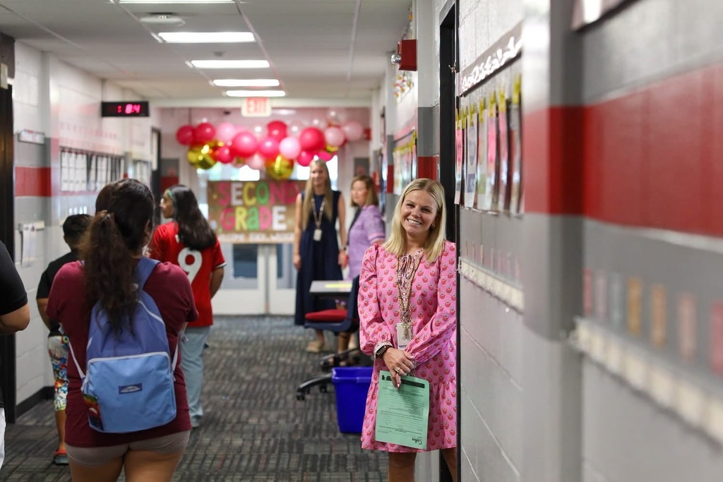 A decorated school hallway lined with red and white walls, filled with teachers and staff, with a pink, red, and gold balloon arch visible in the background.