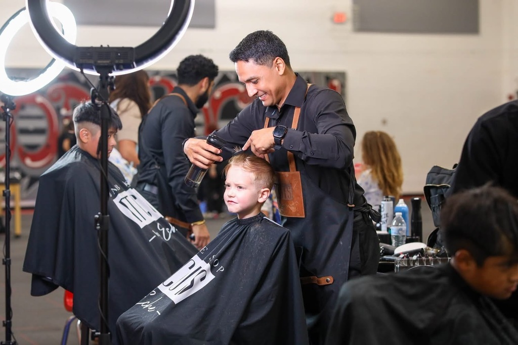 A barber smiling as he cuts a student's hair with clippers during a school event.